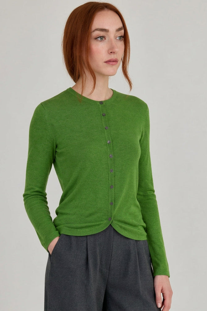 A woman with red hair wearing a bright green long-sleeve button-up cardigan and dark gray pants, standing in a minimalist room with a light gray wall and wooden floor.