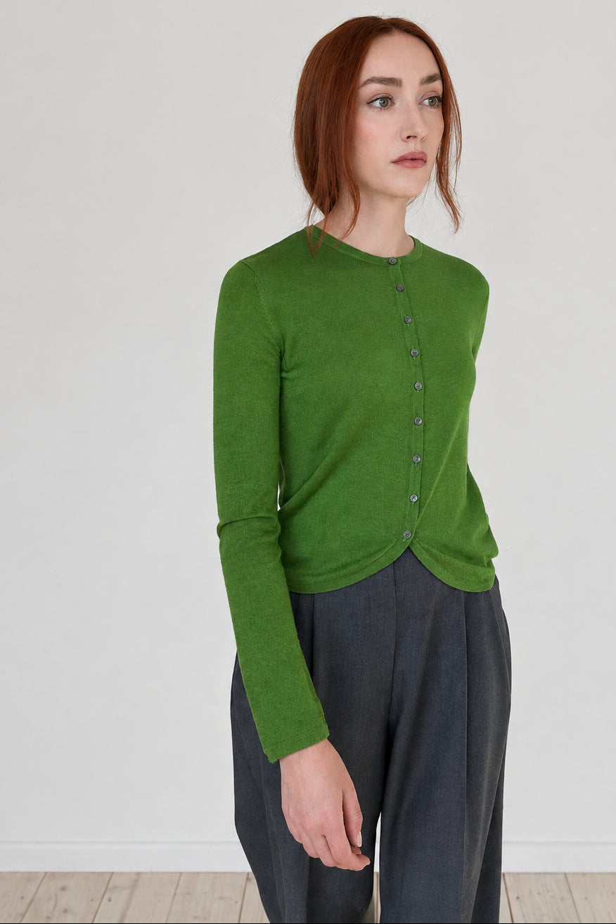 A woman with red hair wearing a bright green long-sleeve button-front cardigan and dark gray wide-leg pants, standing in a minimalist room with a light gray wall and light wood floor.