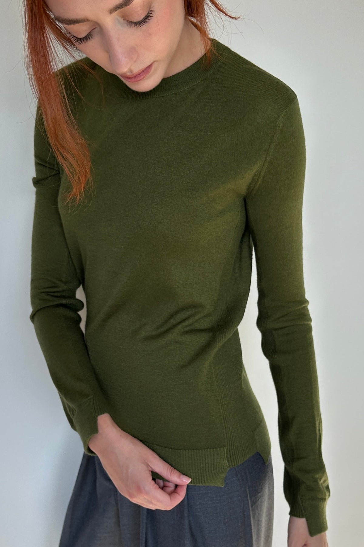 A woman with red hair wearing an olive green long-sleeve mock neck top and gray wide-leg pants, standing in a minimalist room with a white wall and light wood floor.