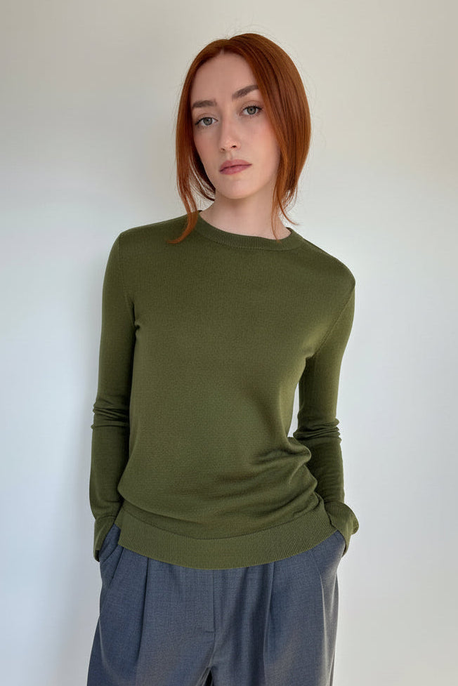 A woman with red hair wearing an olive green long-sleeve knit top and gray wide-leg pants, standing against a plain light gray wall.