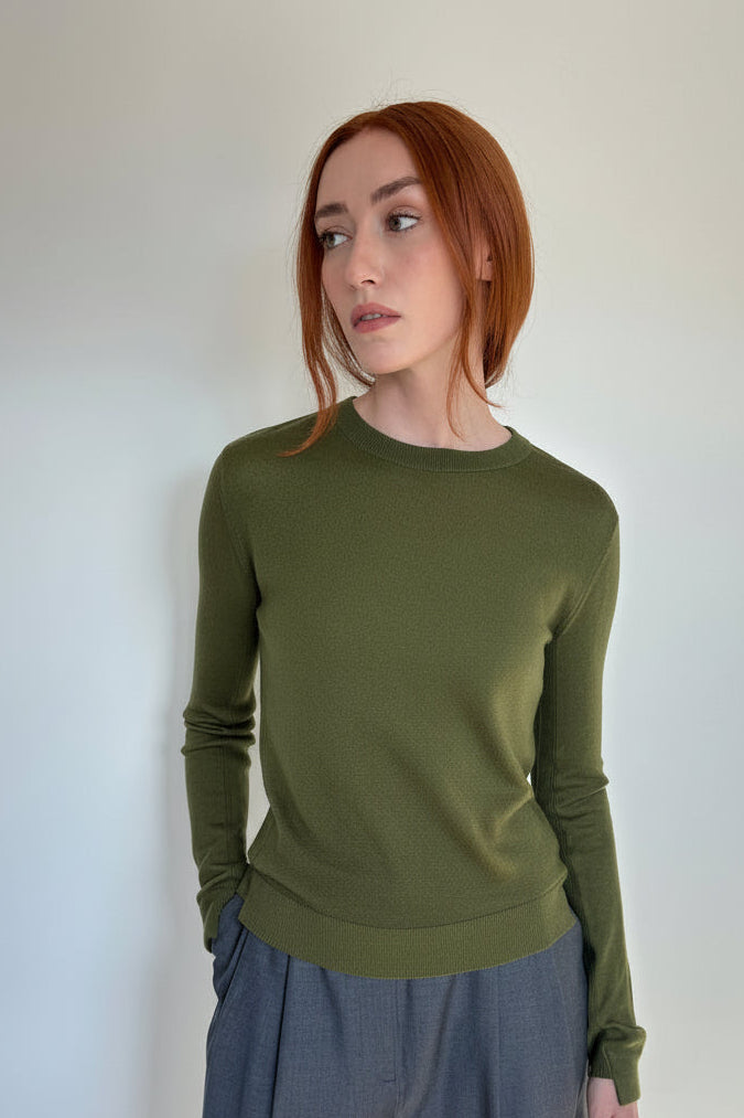 A woman with red hair wearing an olive green long-sleeve knit top and slate blue wide-leg trousers, standing against a plain white wall.