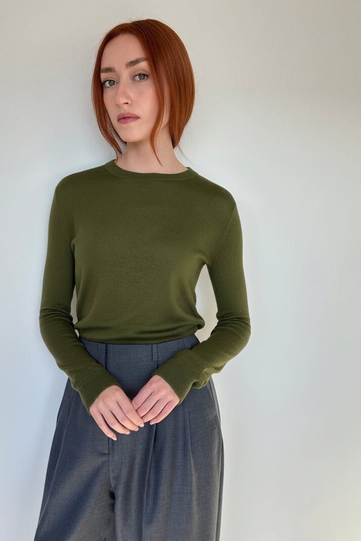 A woman with short red hair stands against a plain light gray wall wearing an olive green long-sleeve knit top and dark gray wide-leg trousers.