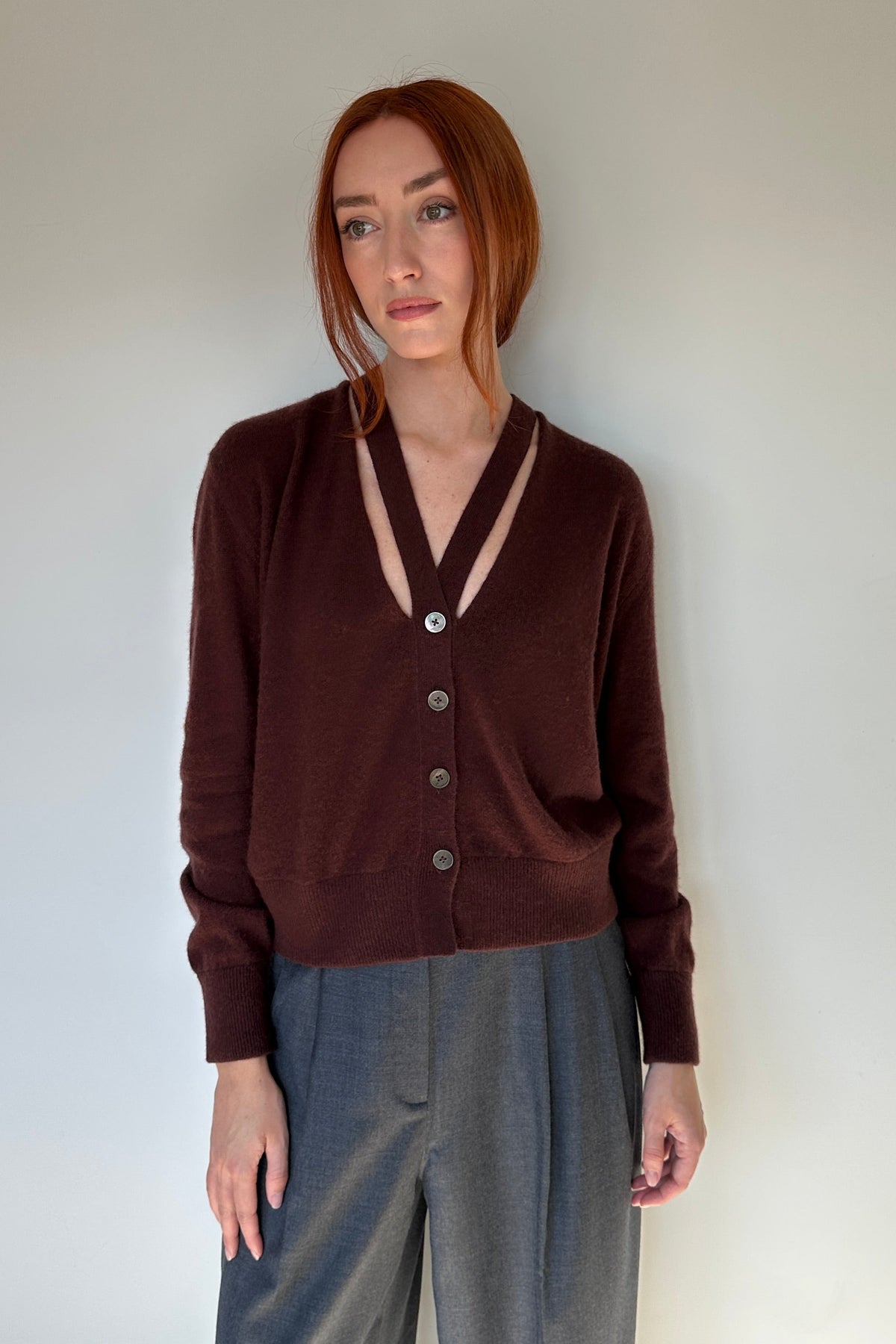 A woman with red hair stands against a plain light gray wall wearing a dark brown button-front cardigan with a V-neck and gray wide-leg pants.