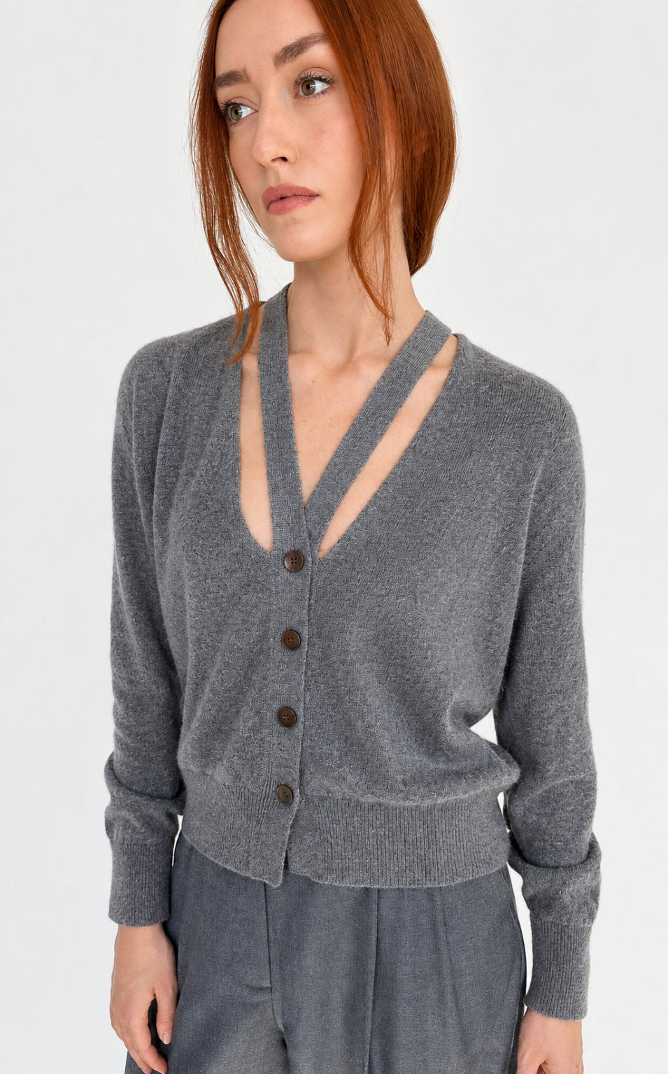 A woman with red hair wearing a brown knit cardigan with a split V-neckline and button-front closure, paired with gray pants, photographed against a plain light background.