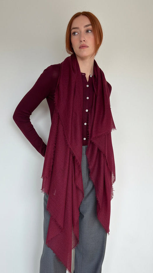 A woman with red hair wearing a long burgundy scarf draped over a button-up shirt and gray pants, standing in a minimalist room with a white wall and light wood floor.