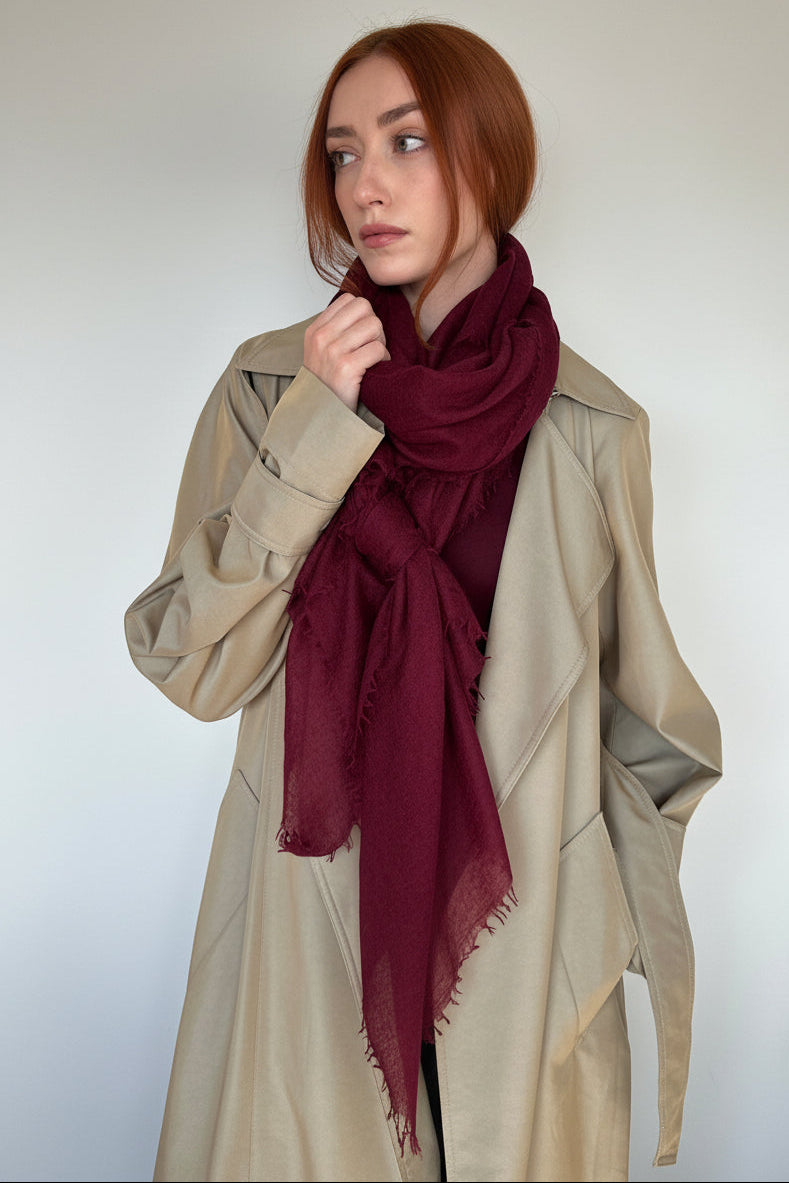 A woman with red hair wearing a beige trench coat and a deep burgundy scarf, posing against a plain off-white wall.