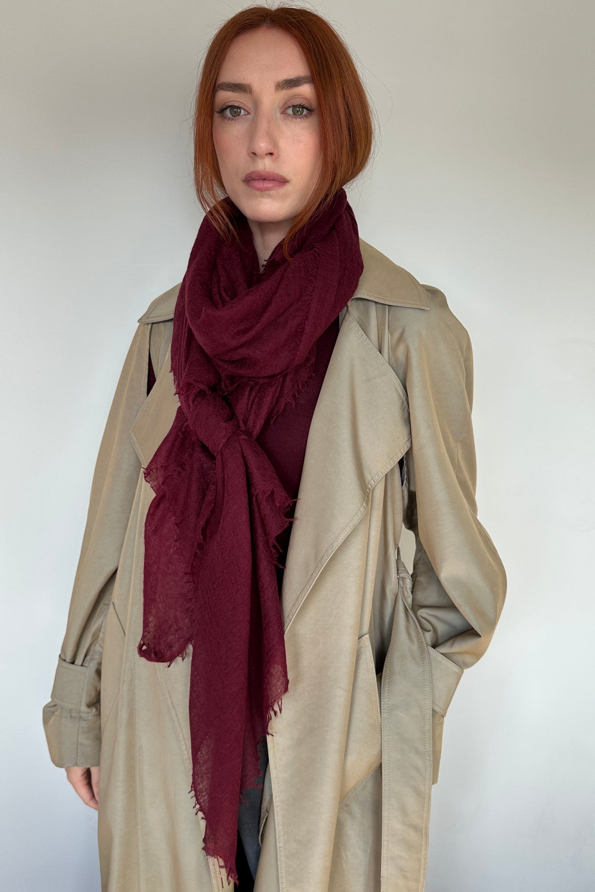 A woman with short red hair wearing a beige trench coat and a deep burgundy scarf, standing against a plain light gray background.