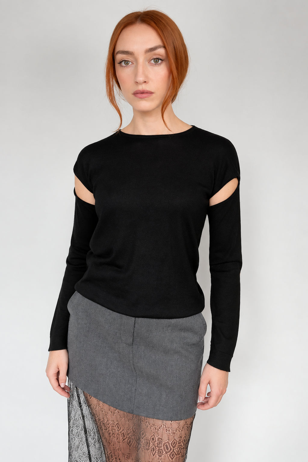 A woman with red hair wearing a black long-sleeve knit top with cutout details on the upper arms, paired with a gray skirt, standing against a plain light gray wall.
