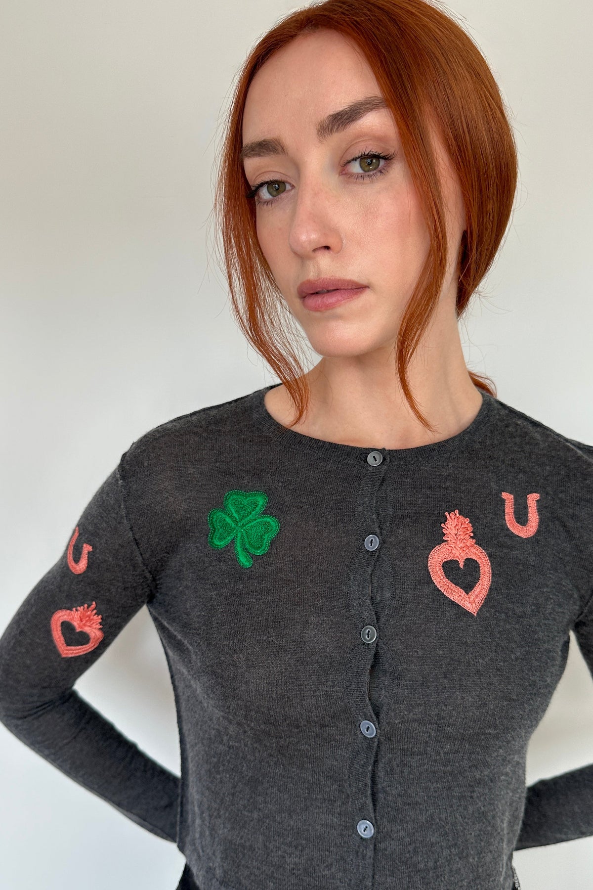A woman with red hair wearing a dark gray long-sleeve button-up cardigan with embroidered St. Patrick's Day motifs, including a green shamrock and pink hearts and letters, against a plain white background.
