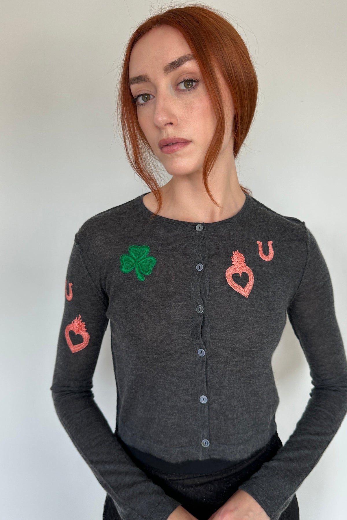 A woman with red hair wearing a dark gray long-sleeve button-up cardigan with embroidered playing card suits (green shamrock, pink hearts, pink horseshoe, pink jack) on a plain light background.