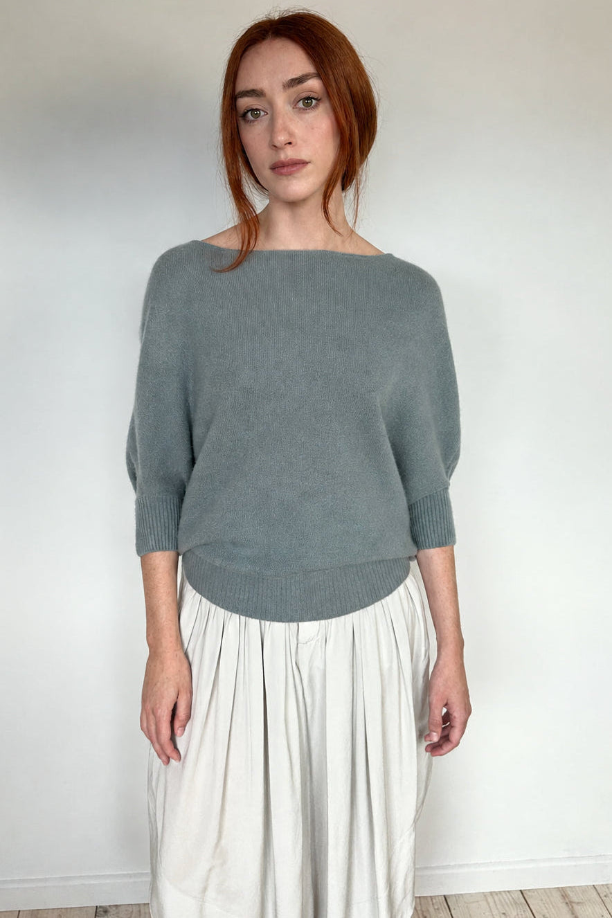 A woman with red hair stands against a plain white wall wearing a muted sage green boat-neck knit sweater and a cream pleated skirt.