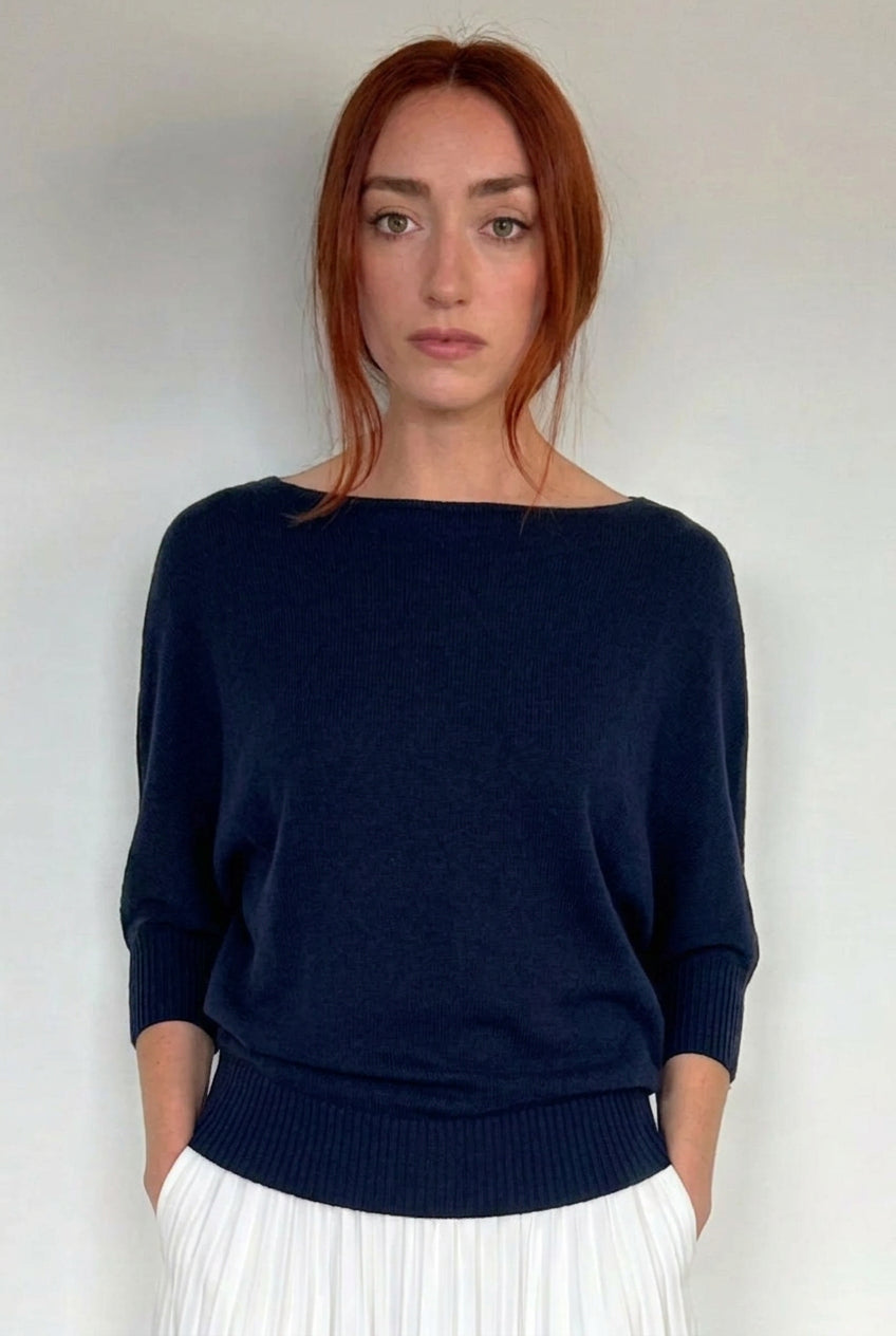 A woman with red hair wearing a navy blue boat-neck knit sweater and a white pleated skirt, standing against a plain light gray background.