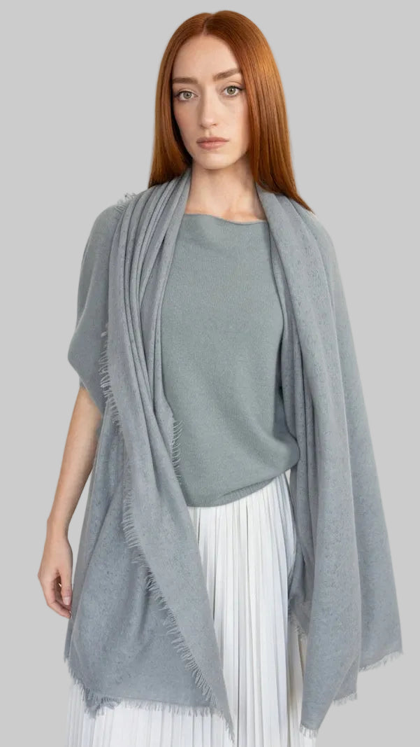 A woman with red hair stands against a white wall wearing a light gray knit top, a white pleated skirt, and a draped light gray shawl with fringed edges.