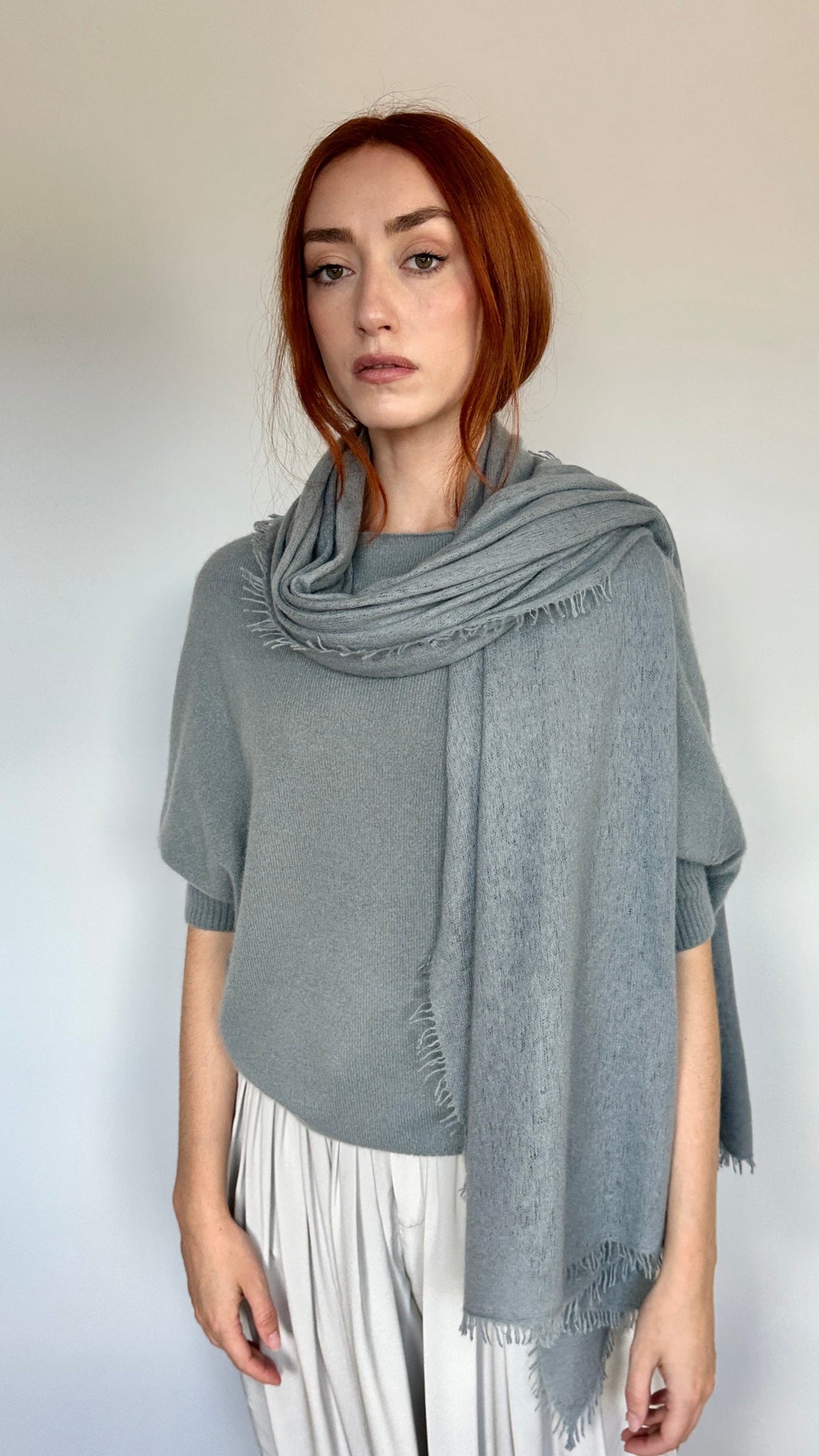 A woman with red hair stands against a plain light wall wearing a draped slate gray knit scarf over a light gray top and white pleated trousers.