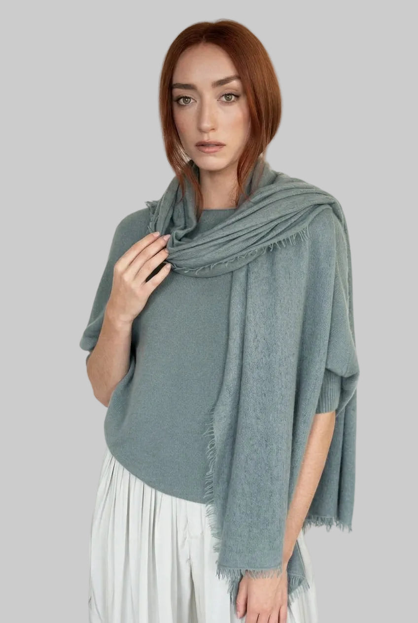 A woman with red hair wearing a sage green knit shawl draped over her shoulders and a white pleated dress, photographed against a plain white studio background.