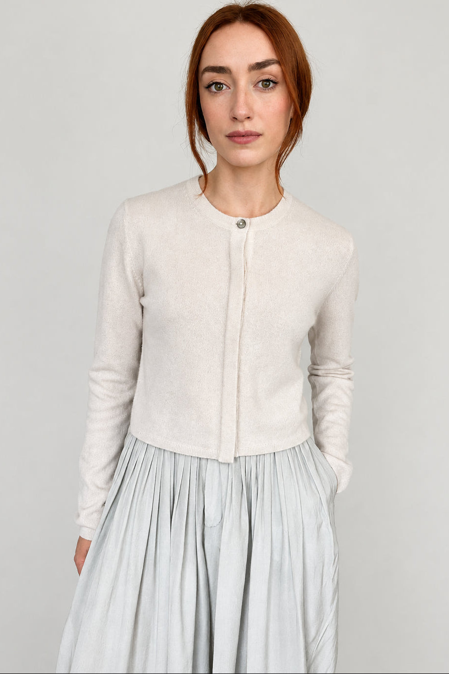 A woman with red hair wearing a cream-colored long-sleeve knit cardigan with a buttoned front and a light gray pleated skirt, standing against a plain light gray wall.