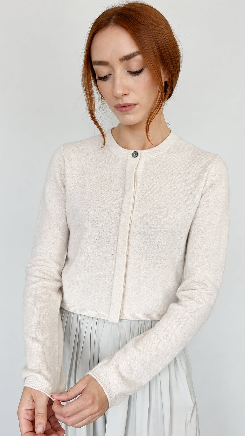 A woman with red hair wearing a cream-colored long-sleeve knit cardigan with a buttoned front over a light gray pleated skirt, standing against a plain light gray wall.