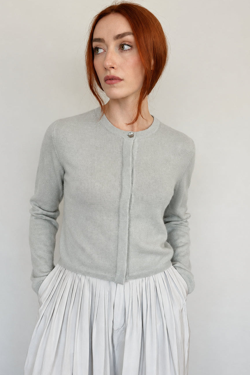 A woman with red hair wearing a light gray knit cardigan with a buttoned crew neck and a pleated light gray skirt, standing against a plain off-white wall.