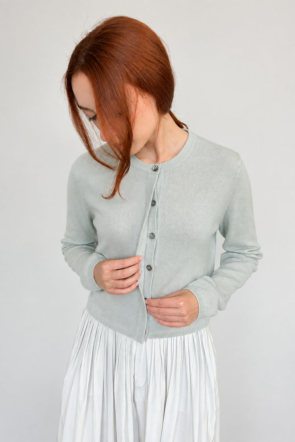 A woman with red hair wearing a light sage green long-sleeve button-up cardigan and a pleated white skirt, posing against a plain off-white wall.