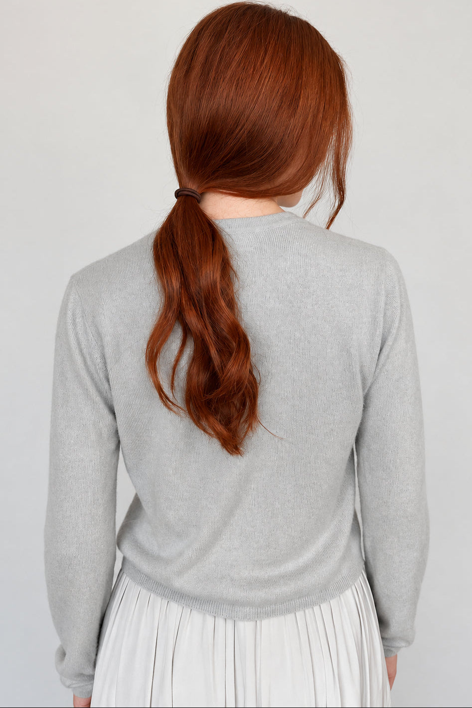 A person viewed from the back wearing a light gray long-sleeve top and a light gray pleated skirt, with auburn hair tied back in a low ponytail against a plain light background.