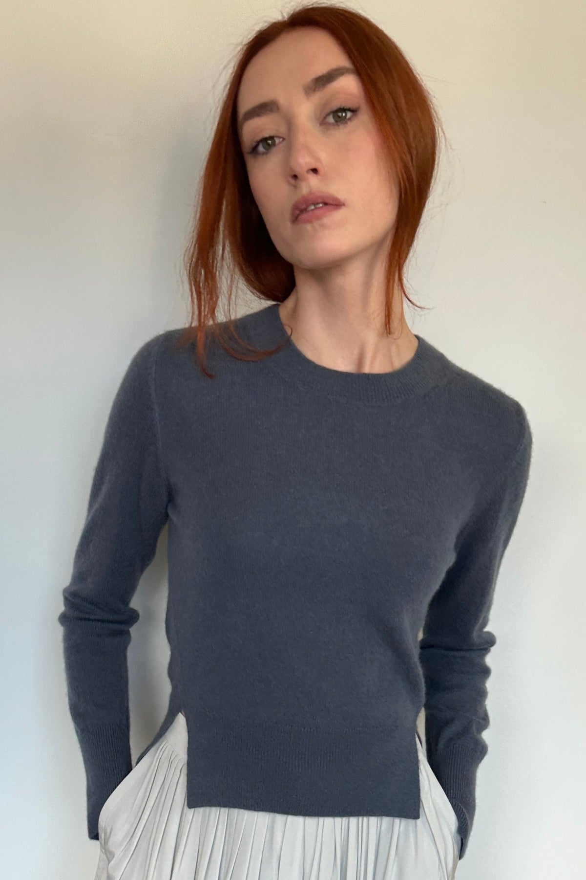 A woman with red hair stands barefoot against a white wall on a light wood floor, wearing a dark charcoal gray long-sleeve knit top and a loose white pleated skirt.