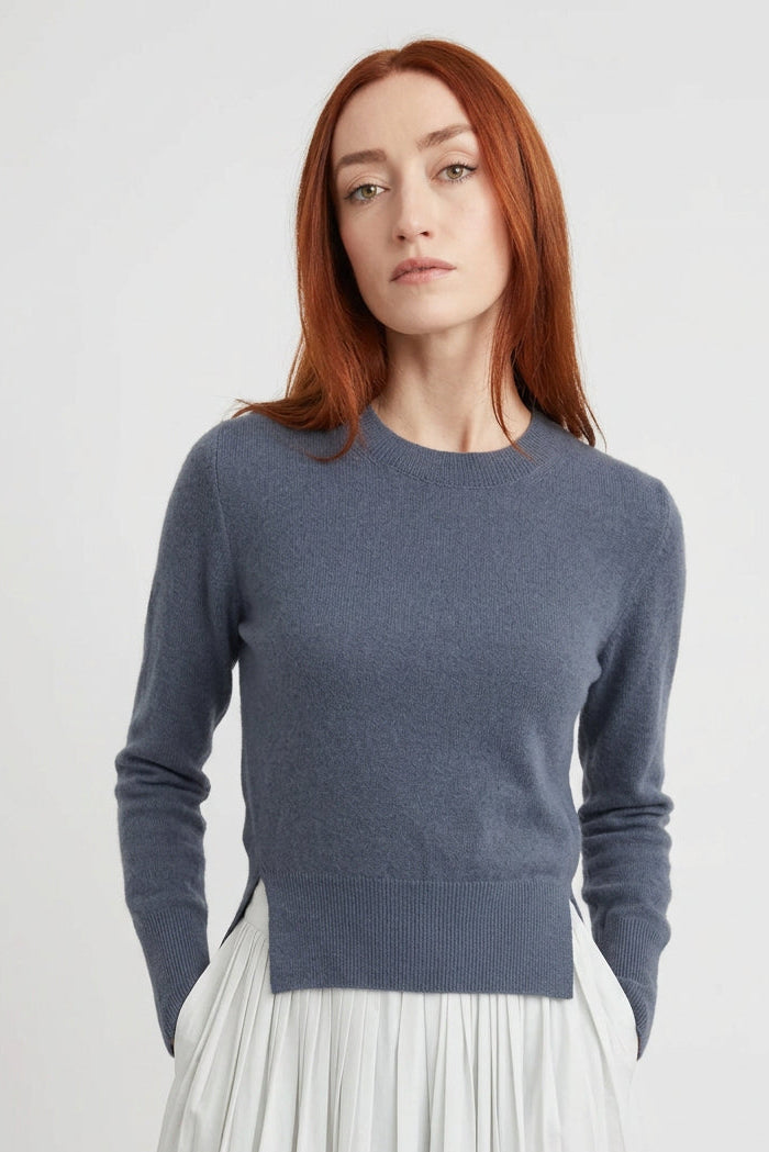 A woman with red hair stands barefoot against a white wall on a light wood floor, wearing a dark charcoal gray long-sleeve knit top and a loose white pleated skirt.