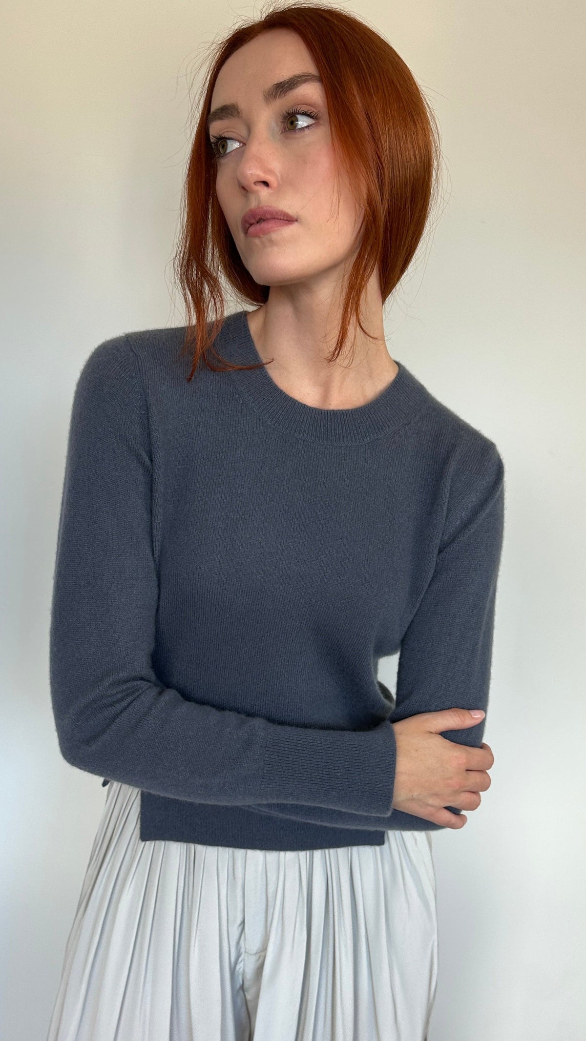 A woman with red hair wearing a dark slate gray long-sleeve crewneck sweater and light beige pleated trousers, standing against a plain off-white wall.