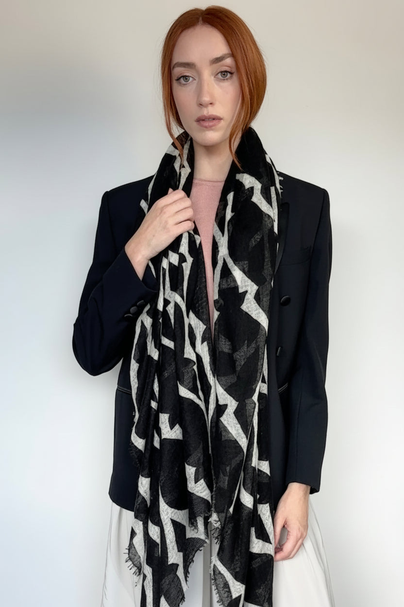 A woman with red hair wearing a black and white abstract patterned scarf, a black blazer, and a light pink top, standing against a plain light gray wall.