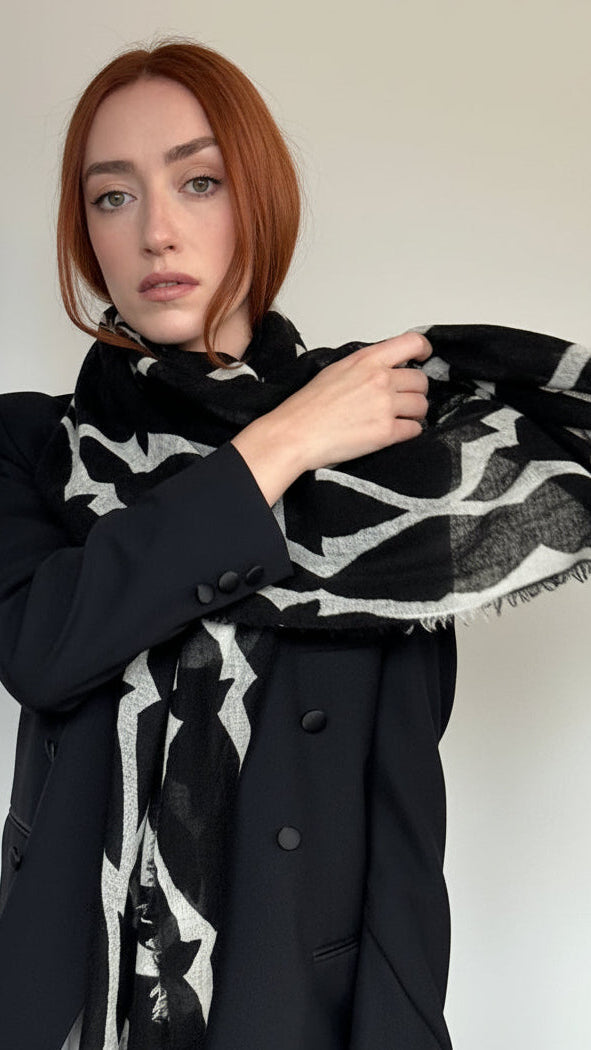 A woman with red hair wearing a black coat and a black-and-white patterned scarf, holding the scarf out to show its pattern against a plain light background.