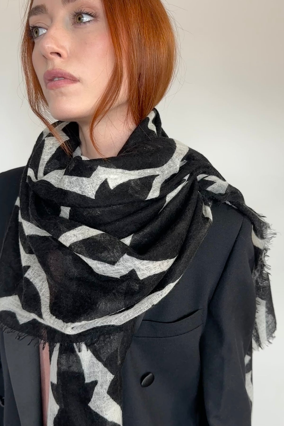 A close-up of a woman with red hair wearing a black and light gray patterned scarf over a black blazer, set against a plain light background.