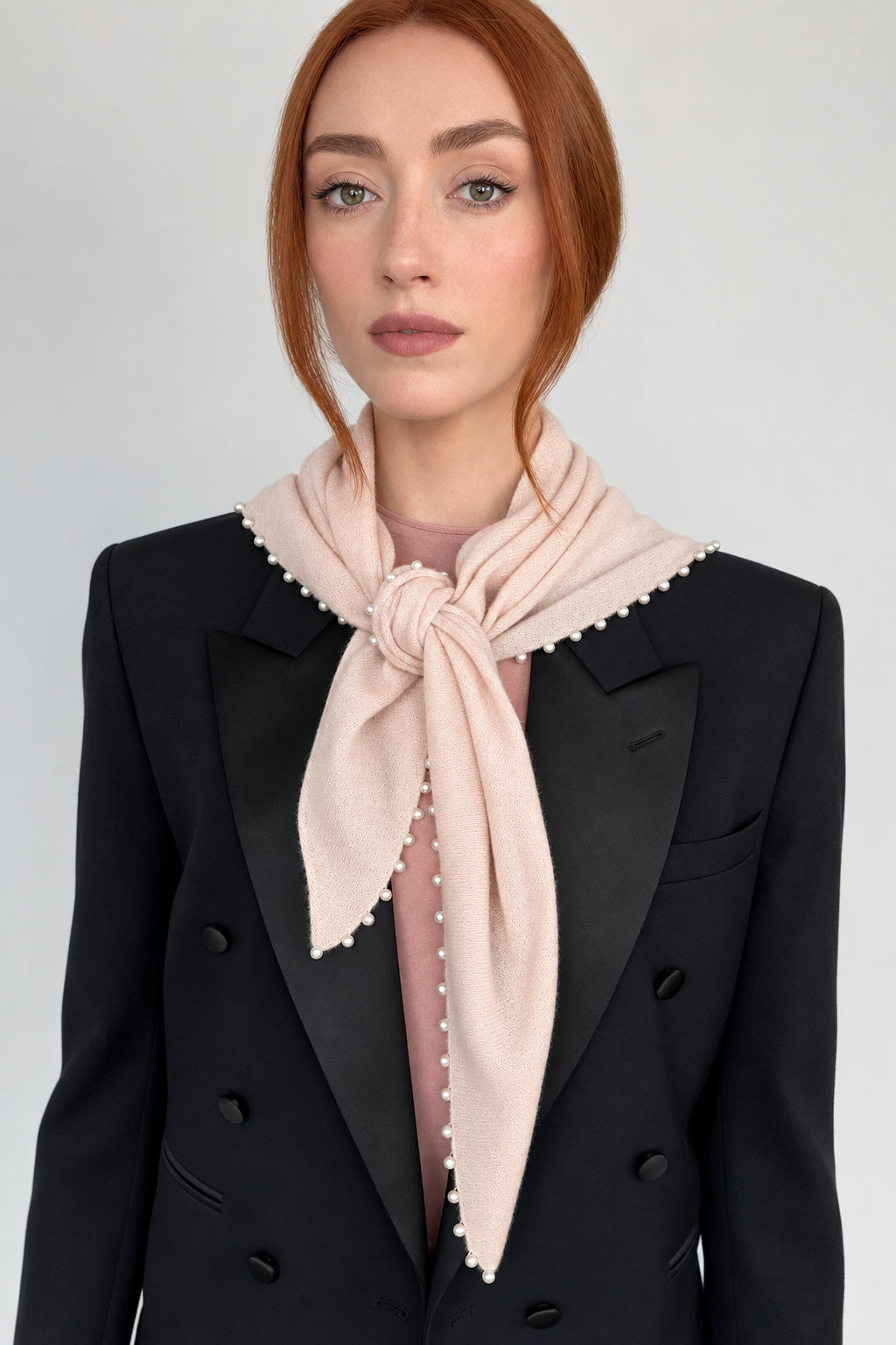 A woman with red hair wearing a black double-breasted blazer and a light pink knotted scarf with white pearl trim, set against a plain light gray background.