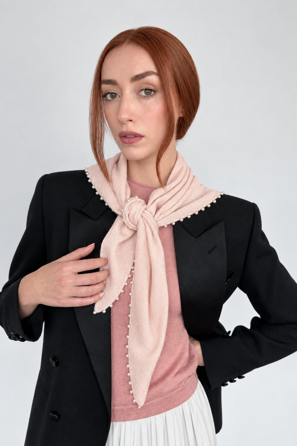 A woman with red hair wearing a black blazer, a dusty pink knit top, and a light pink scarf with white pearl edges tied around her neck, set against a plain off-white background.
