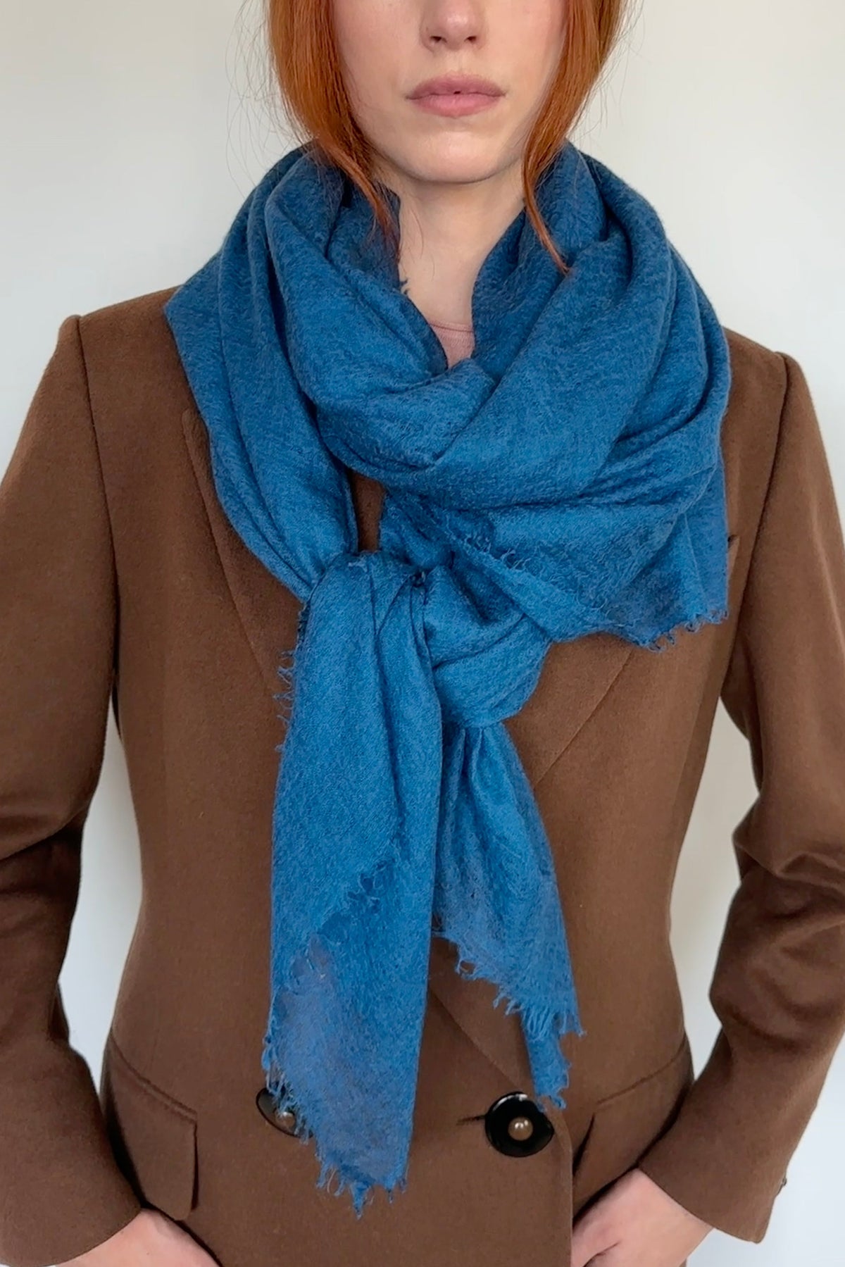 A woman with red hair wearing a brown blazer and a bright teal petrol blue scarf, photographed against a plain white background.