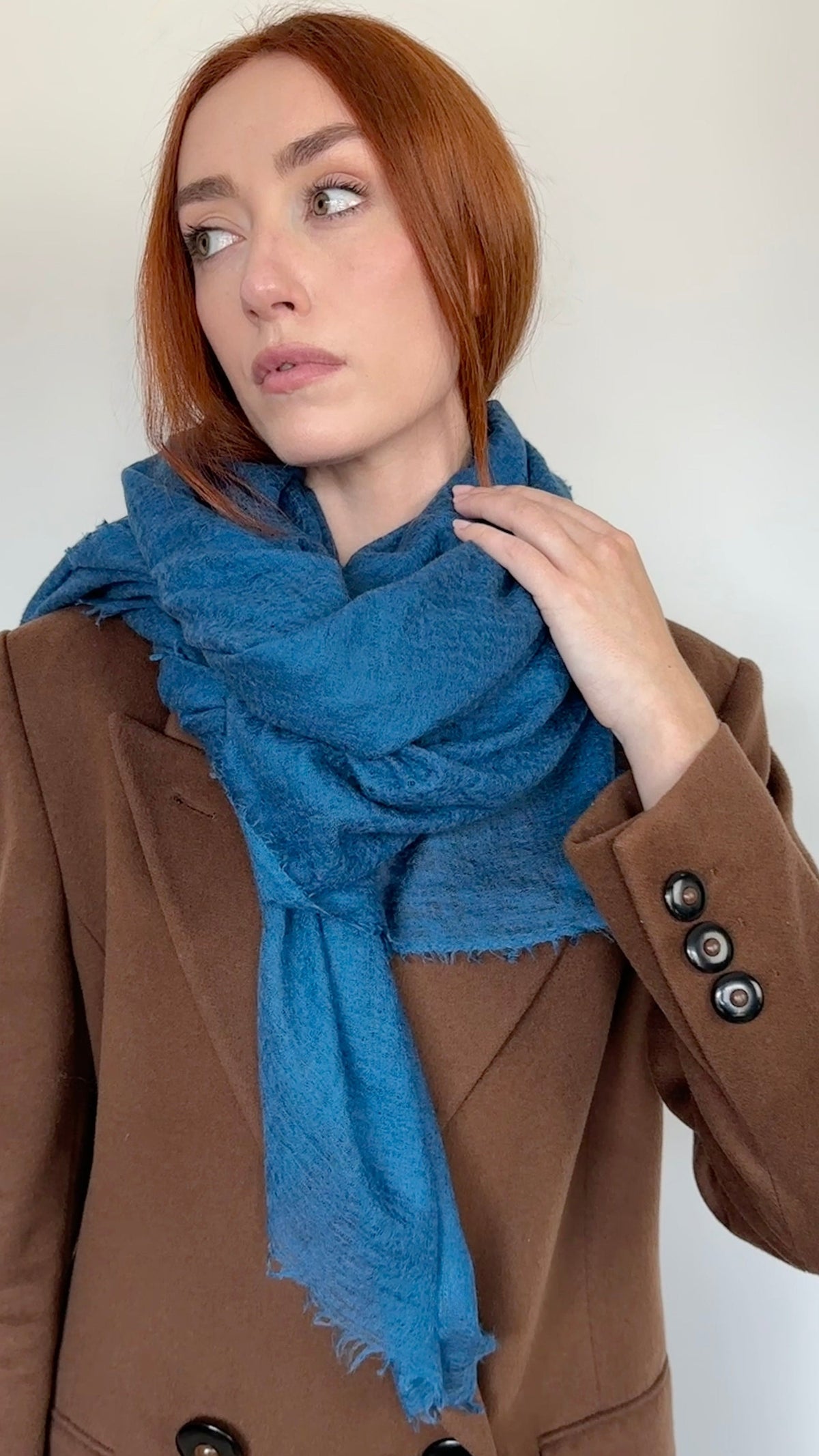 A woman with red hair wearing a brown double-breasted coat and a cashmere teal petrol blue scarf, posing against a plain off-white wall.