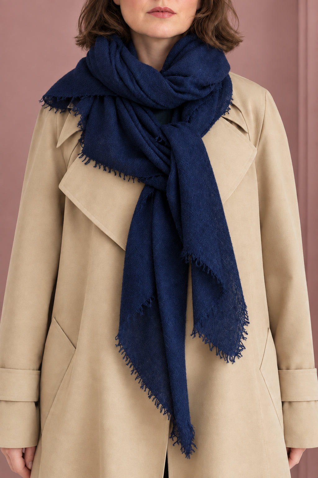 A woman wearing a beige trench coat dark navy blue scarf, standing in front of a pink door frame.