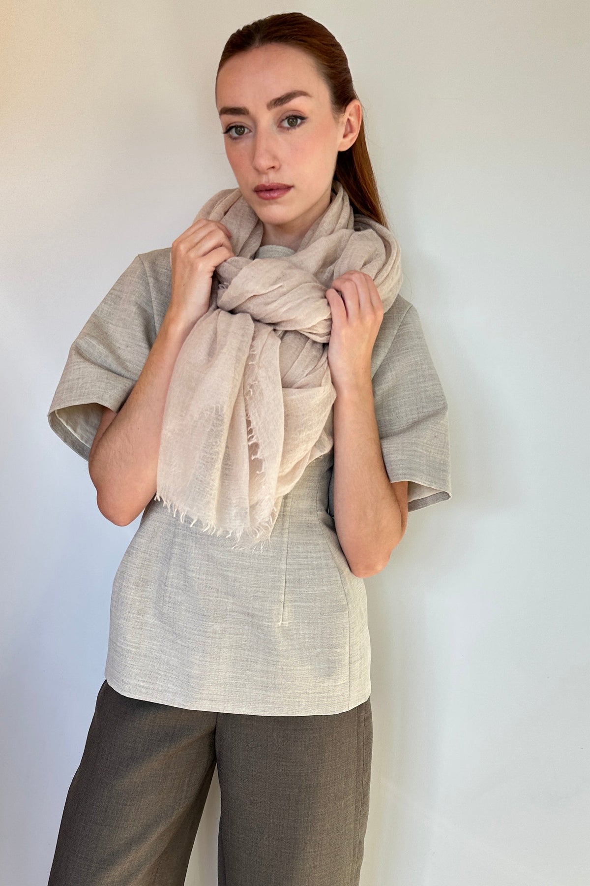 A woman modeling a light beige cashemere scarf wrapped around her neck, paired with a light gray short-sleeve top and dark gray trousers, against a plain off-white wall.