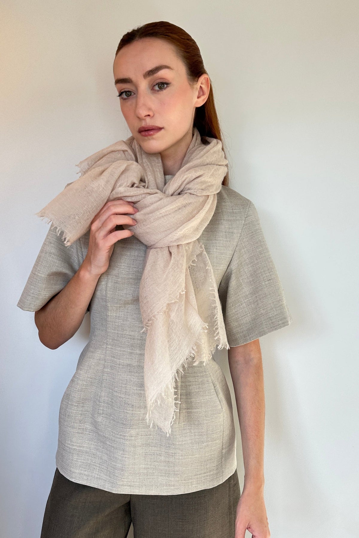 A woman modeling a light beige cashmere fringed scarf wrapped around her neck, paired with a textured light gray short-sleeve top and dark gray pants, standing against a plain off-white wall.
