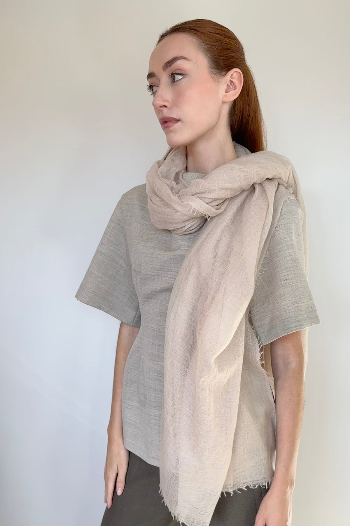 A woman wearing a light beige cashmere scarf draped over her shoulders, paired with a gray short-sleeve top and olive green pants, standing against a plain white wall.
