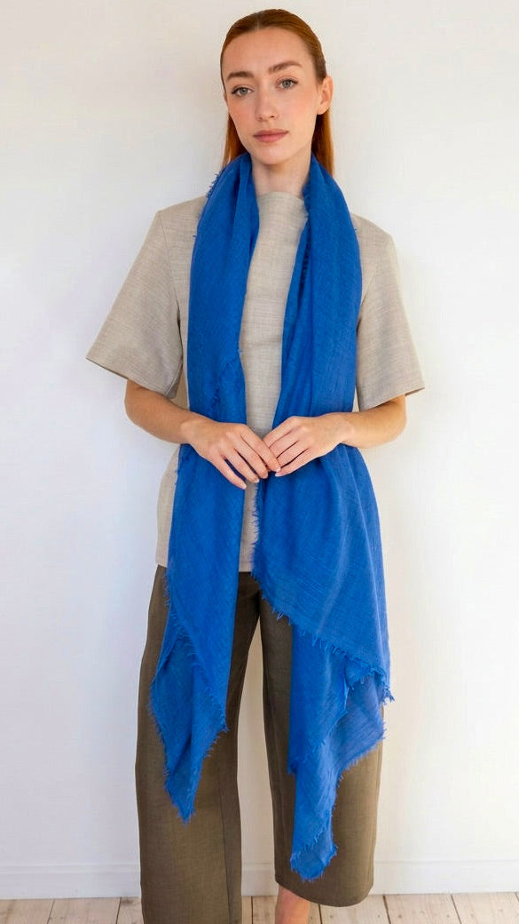A woman modeling a bright blue fringed scarf wrapped around her neck, paired with a light gray t-shirt and brown pants, standing against a plain white wall.
