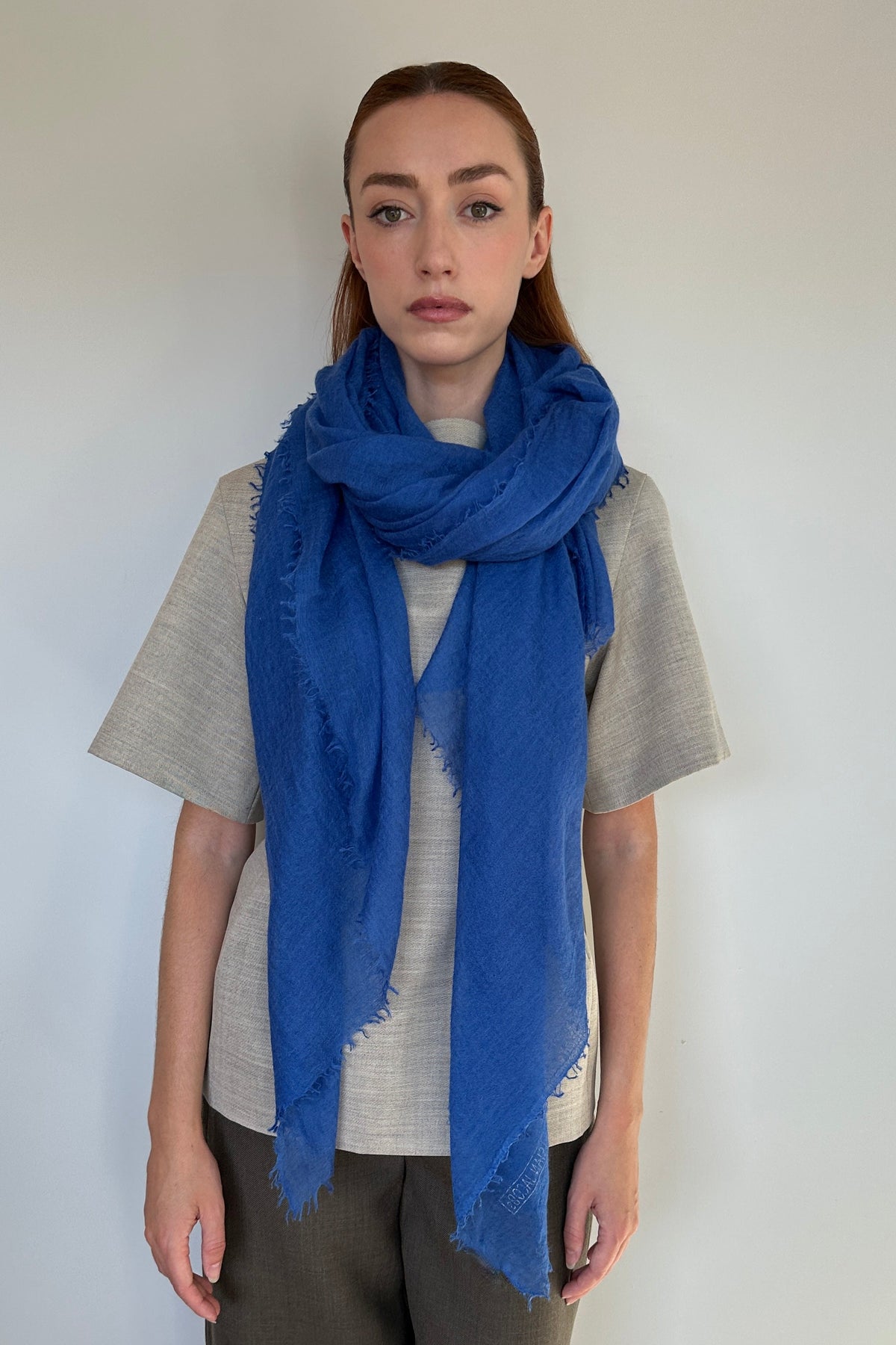 A woman modeling a vibrant royal blue scarf with fringed edges, wrapped around her neck over a light gray t-shirt and dark brown trousers, standing against a plain off-white wall.