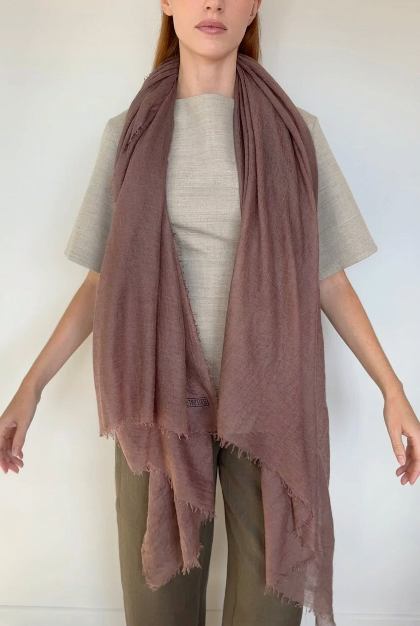 A woman wearing a draped dusty taupe mauve-brown scarf over a light gray top and olive pants, standing against a plain white wall.