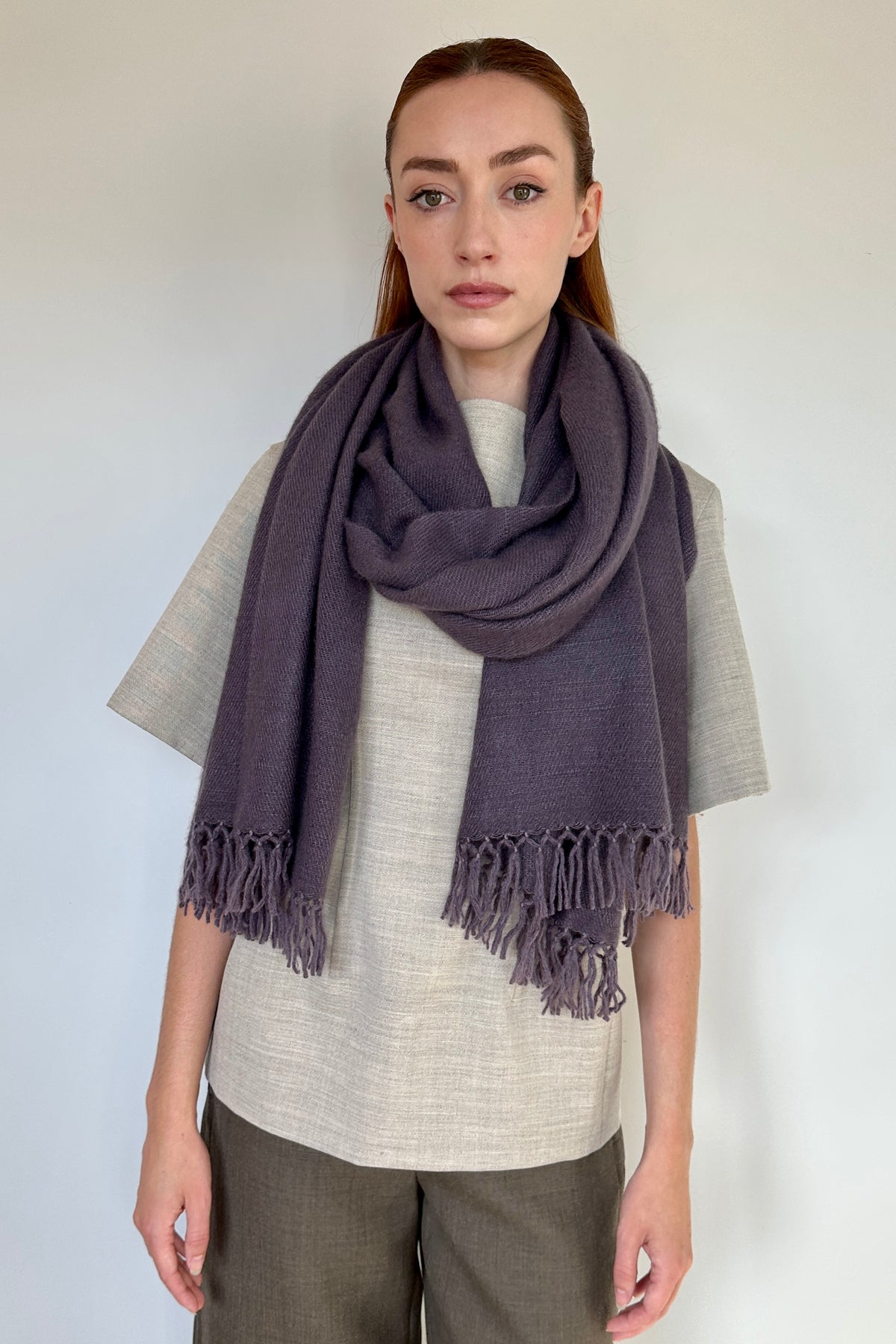 A woman modeling a dark purple cashmere fringed scarf wrapped around her neck, paired with a light beige linen shirt and brown trousers, against a plain white wall.