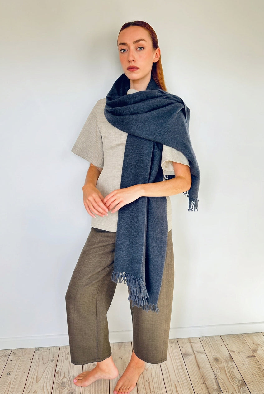 A woman stands barefoot in a minimalist room with light wood floors and a white wall, wearing a large navy blue fringed scarf or shawl draped over her shoulders, a light gray short-sleeve top, and olive-brown cropped pants.
