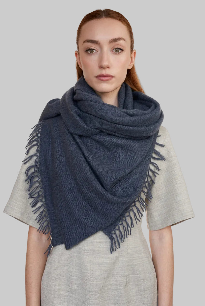 A woman modeling a large dusty navy cashmere fringed shawl scarf draped over her shoulders, paired with a light beige linen dress against a plain light gray background.