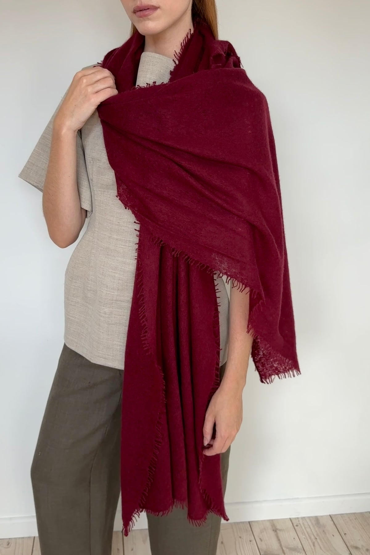 A person wearing a large deep burgundy cashmere knit shawl with fringed edges over a light beige top and olive green pants, standing against a plain white wall.