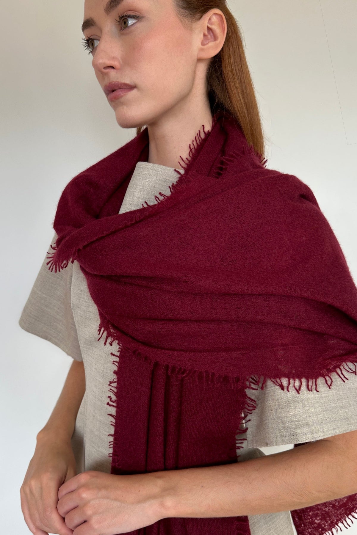 A woman wearing a deep burgundy cashmere knitfringed shawl or wrap over a light beige linen top, photographed against a plain off-white background.