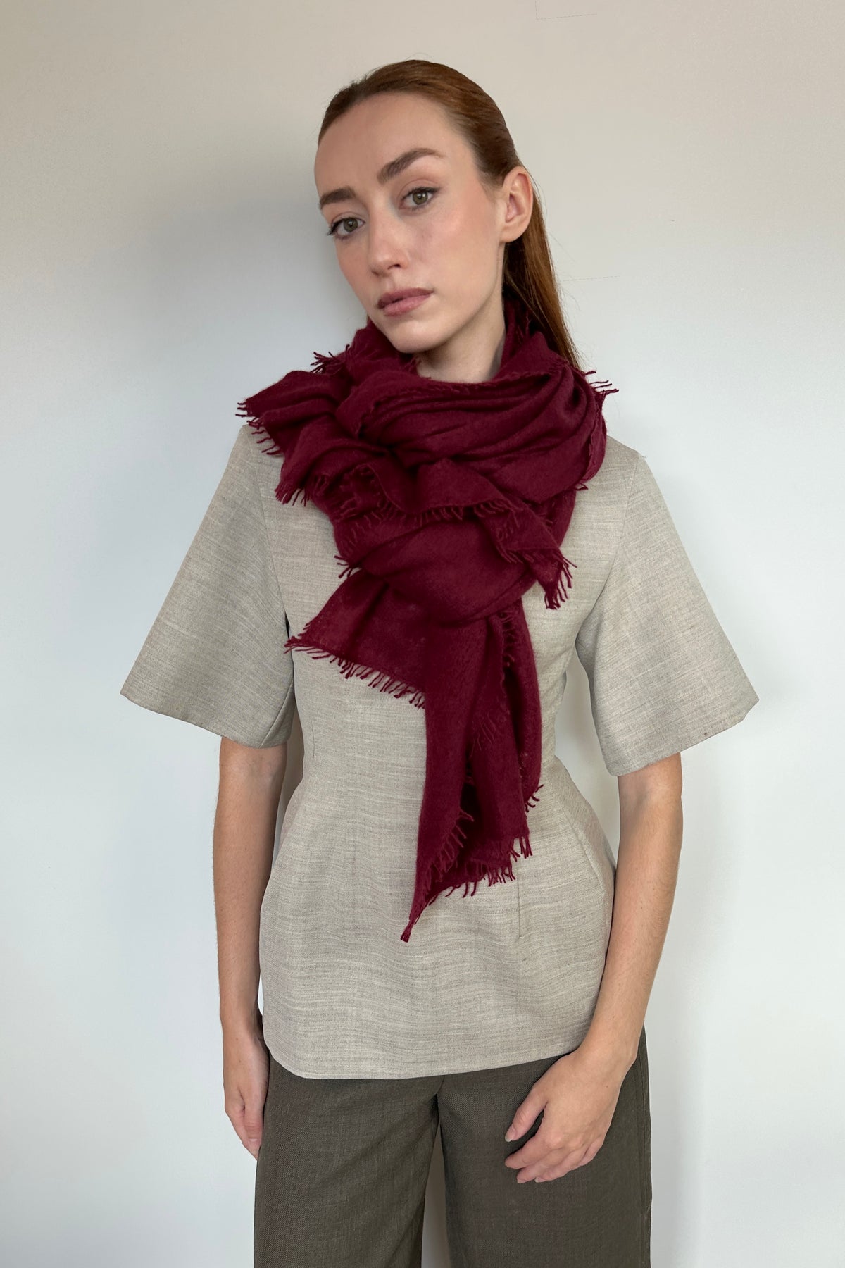 A woman modeling a deep burgundy cashmere knit fringed scarf wrapped around her neck, paired with a light beige linen short-sleeve top and dark gray trousers, against a plain light gray background.