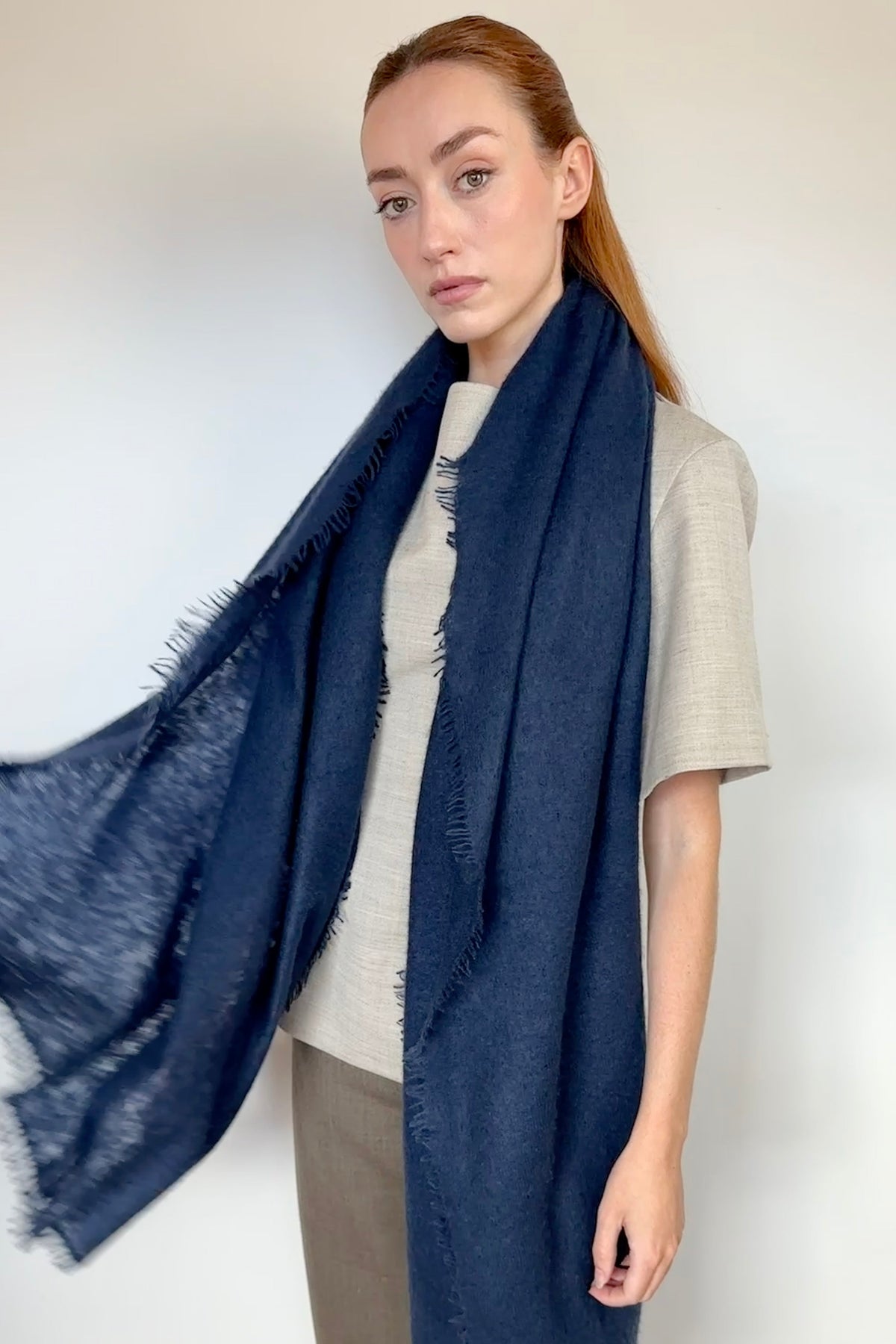 A woman modeling a large navy blue cashmere fringed scarf draped over her shoulders, paired with a beige top and brown pants against a plain white wall.
