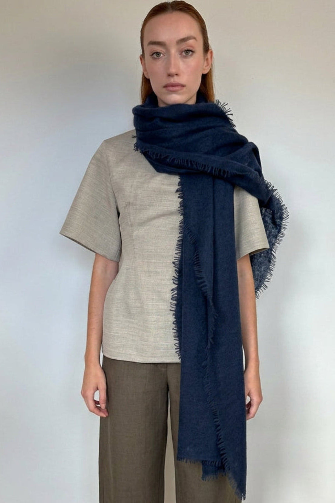 A woman modeling a dark navy blue fringed shawl or wrap draped over her shoulders, paired with a light beige short-sleeve top and brown cropped pants, standing against a plain white wall.