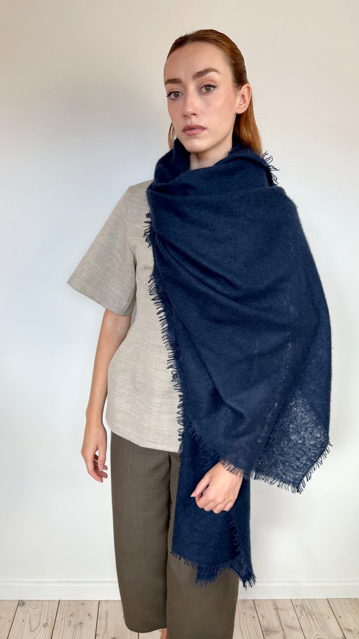 A woman modeling a large navy blue cashmere shawl with fringed edges, draped over her shoulders, paired with a light beige short-sleeve top and brown trousers against a plain white wall.